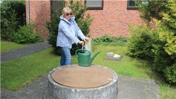 Eine Frau in Jeans und hellblauer Jacke füllt an einer Wasserstelle auf dem Friedhof Wasser in ihre Gießkanne