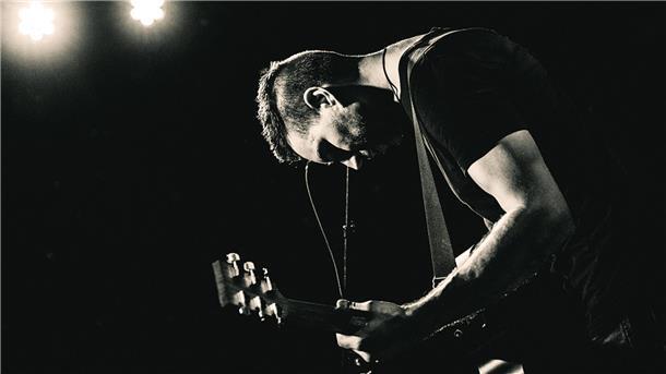 Musiker Troy Petty mit Gitarre auf einem Schwarz-Weiß-Foto im Scheinwerferlicht.