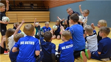 Ein Mann sitzt mit vielen Kindern im Kreis auf dem Boden einer Sporthalle. Ein paar der Kinder haben einen Fußball in der Hand oder melden sich für eine Wortmeldung. 