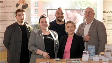 Fünf Personen – ein Mann, eine Frau, ein Mann, eine Frau und ein Mann - stehen an einem Tisch. Sie gucken in die Kamera.