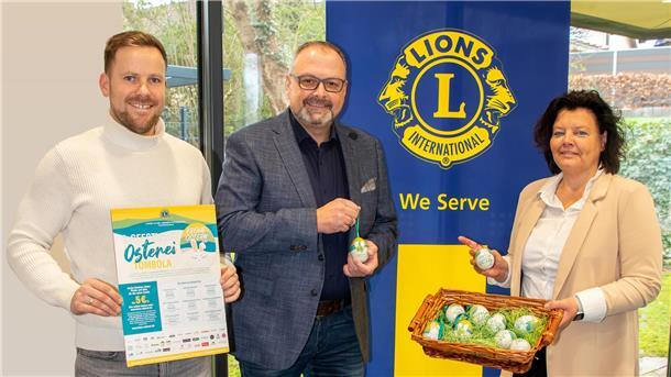 Drei Personen stehen vor einem Banner mit dem Logo des Lions Clubs International. Der Mann links hält ein Plakat zur „6. Geestland Osterei-Tombola“, der Mann in der Mitte zeigt ein Osterei mit Aufdruck, und die Frau rechts hält einen Korb mit weiteren dekorativen Ostereiern.