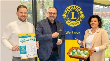 Drei Personen stehen vor einem Banner mit dem Logo des Lions Clubs International. Der Mann links hält ein Plakat zur „6. Geestland Osterei-Tombola“, der Mann in der Mitte zeigt ein Osterei mit Aufdruck, und die Frau rechts hält einen Korb mit weiteren dekorativen Ostereiern.