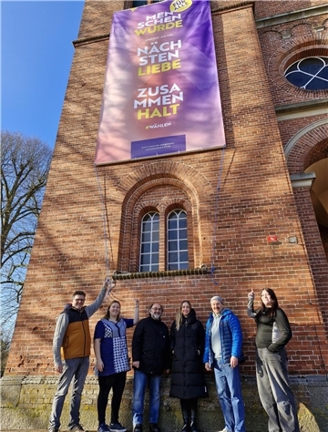 Eine Gruppe von sechs Personen steht vor einer Backsteinfassade mit einem großen lilafarbenen Banner, das an der Wand hängt. Auf dem Banner stehen die Worte „Menschenwürde“, „Nächstenliebe“ und „Zusammenhalt“ in gelber und weißer Schrift. Die Gruppe, bestehend aus Männern und Frauen, lächelt und zeigt mit den Händen auf das Banner. 