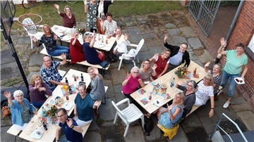 Eine Gruppe von Menschen sitzt an Tischen und winken in die Kamera.