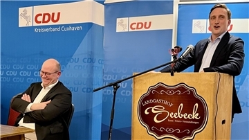 Ein Mann sitzt mit verschränkten Armen am Tisch und lacht, ein weiterer steht am Rednerpult. Hinter ihnen Banner der CDU.