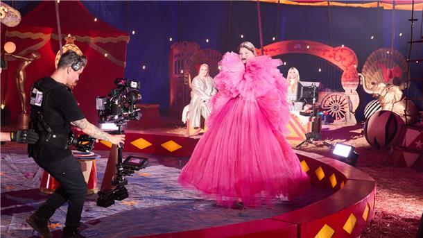 Frau trägt ein pinkes, Pompöses Kleid in einem Zirkuszelt, sie läuft in der Manege. Ein Mann mit Kamera geht rückwärts vor ihr her. Im Hintergrund sind zwei blonde Frauen zu sehen.