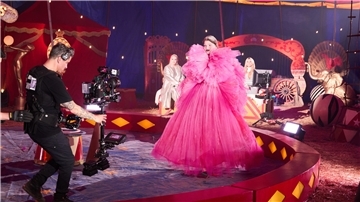 Frau trägt ein pinkes, Pompöses Kleid in einem Zirkuszelt, sie läuft in der Manege. Ein Mann mit Kamera geht rückwärts vor ihr her. Im Hintergrund sind zwei blonde Frauen zu sehen.
