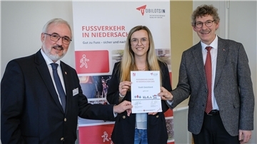 **ALT-Text:**  
Drei Personen bei der Urkundenübergabe zum Fußverkehrs-Check Niedersachsen; eine Frau in der Mitte hält die Urkunde der Stadt Geestland, flankiert von zwei Männern im Anzug, im Hintergrund ein Roll-up mit der Aufschrift „Fußverkehr in Niedersachsen“.