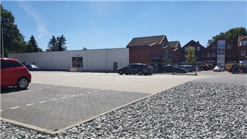 Ein Supermarktparkplatz, auf dem Autos parken, wird von einer Schotterfläche umrandet.