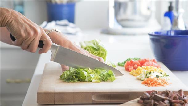 Meal Prep für Berufstätige: Gesund essen und Zeit sparen ist keine Zauberei Auf einem Holzbrett wird Salat mit einem großen Messer geschnitten. Im Hintergrund stehen Küchenutensilien.