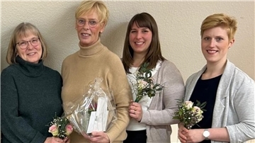 Vier Frauen stehen nebeneinander vor einer beigen Wand und lächeln in die Kamera. Die Frauen halten kleine Blumensträuße mit rosa und weißen Blüten, während die zweite Frau von links zusätzlich ein Geschenk in transparenter Verpackung trägt.