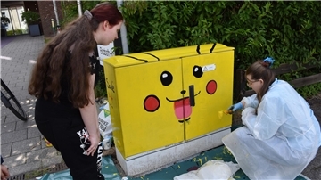 Zwei Schülerinnen stehen an einem Stromkasten, den sie leuchtend gelb wie die Pokemon-Figur Pikachu angemalt haben.