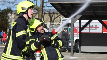 Zwei angehende Feuerwehrleute halten einen wassersprühenden Schlauch fest. Im Hintergrund steht ein Feuerwehrwagen der Bramstedter Feuerwehr. 