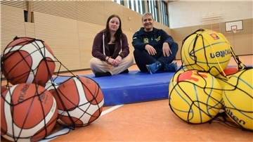 Eine junge Frau und ein Mann sitzen in einer Sporthalle auf einer Matte, davor liegen Bälle.