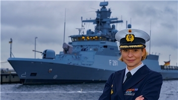 Bianca Seifert blickt in die Kamera. Im Hintergrund liegt eine Korvette vor Anker.