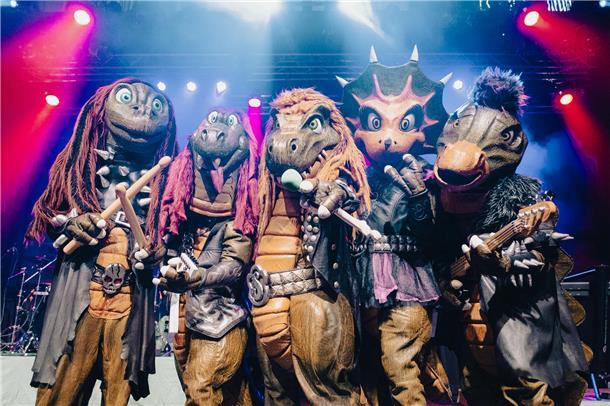 Fünf als Dinosaurier verkleidete Menschen stehen auf einer Bühne. Heavysaurus liefern handgemachten Rock und Metal mit kindgerechten Texten: Die grüngeschuppten Urzeitwesen stehen am 25. April um 17.30 Uhr auf der Bühne in der Stadthalle Bremerhaven. 