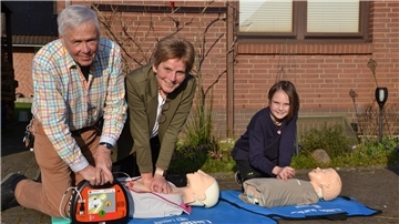 Ein Mann und eine Frau simulieren die Herzdruckmassage an einer Puppe, der Mann hält einen Defibrillator bereit. Neben ihnen simuliert ein Mädchen die Herzdruckmassage an einer Puppe.