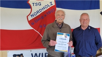 Vor einer Flagge der TSG Nordholz hält Heinz-Dieter Kuhl seine Urkunde für 65 Jahre Vereinszugehörigkeit. Neben ihm steht der Vorsitzende Frank Bretschneider.