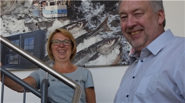 Ein Mann und eine Frau stehen auf einer Treppe. Im Hintergrund hängt ein Bild, auf dem Fische und Eis gemalt sind.