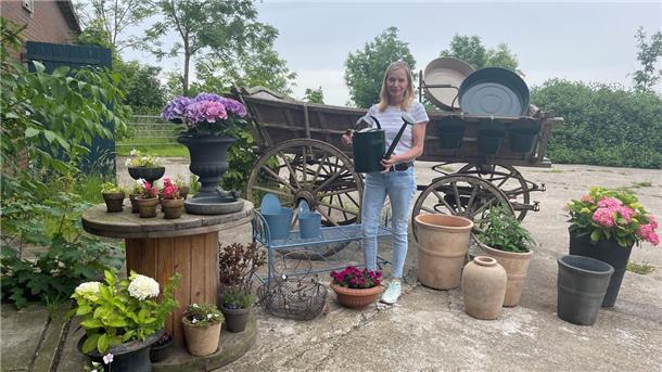 Frau mit Gießkanne vor einem alten Holzwagen, rundherum Blumentöpfe.