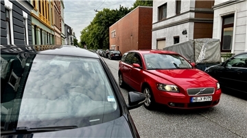 Man sieht ein rotes Auto in einer kleinen Straße. Am Rand parken überall Autos.