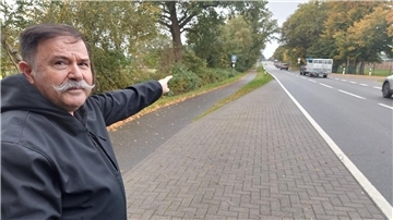 Ein Mann mit einem langen Schnurrbart zeigt mit der Hand auf eine Landstraße. 