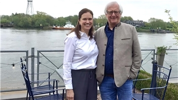 **ALT-Text:**  
Ilka Studnik und Karl-Ernst Thümler stehen lächelnd nebeneinander an einem Geländer mit Blick auf einen Fluss; im Hintergrund eine Fähre und grüne Uferlandschaft.