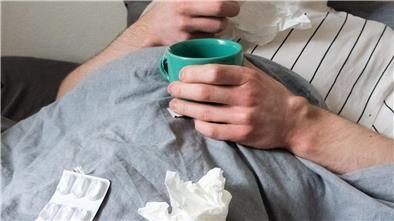 ILLUSTRATION: Ein Mann liegt mit Taschentüchern, Teetasse, Nasenspray und Tabletten im Bett.