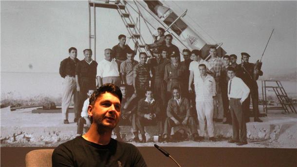 Ein Mann sitzt vor einem Mikrofon auf einer Bühne, hinter ihm ist ein historisches Schwarz-Weiß-Foto projiziert, das eine Gruppe von Männern vor einer Rakete zeigt.