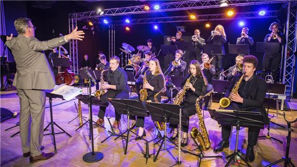 Dirigent in grauem Anzug leitet ein Jazzorchester mit Bläsern und Schlagzeug auf einer beleuchteten Bühne. Die Musiker spielen konzentriert ihre Instrumente, während bunte Scheinwerfer die Szene in warmen Farben beleuchten.