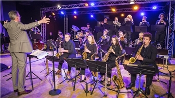Dirigent in grauem Anzug leitet ein Jazzorchester mit Bläsern und Schlagzeug auf einer beleuchteten Bühne. Die Musiker spielen konzentriert ihre Instrumente, während bunte Scheinwerfer die Szene in warmen Farben beleuchten.