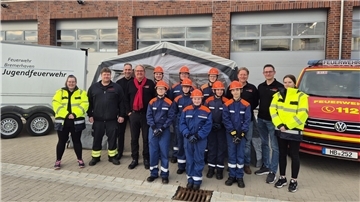 Gruppenfoto vor einer Feuerwehrwache mit Mitgliedern der Jugendfeuerwehr Bremerhaven und Betreuenden. Im Hintergrund sind ein neues Zelt, ein Feuerwehranhänger mit der Aufschrift „Jugendfeuerwehr“ sowie ein Einsatzfahrzeug zu sehen. Die Jugendlichen tragen blaue Schutzanzüge mit orangefarbenen Helmen.