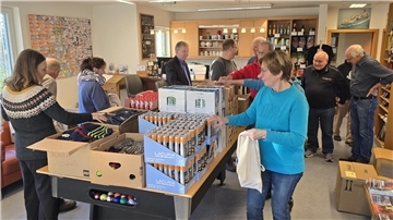 Mehrere Personen stehen in einem hellen Raum und sortieren Produkte wie Duschgel, Schals und andere Artikel auf einem Tisch. Die Menschen wirken beschäftigt.