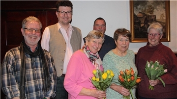 Gruppenfoto von sechs älteren Erwachsenen, drei Männer und drei Frauen, die in einem Raum mit Holzwänden und einem gerahmten Gemälde im Hintergrund stehen. Drei der Frauen halten bunte Blumensträuße in den Händen.