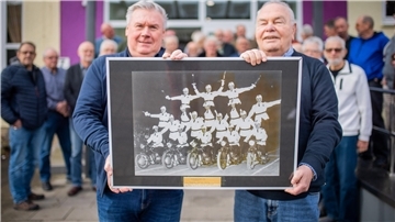 Zwei Männer halten ein Foto in die Kamera. Das Bild zeigt eine menschliche Pyramide auf Motorrädern.