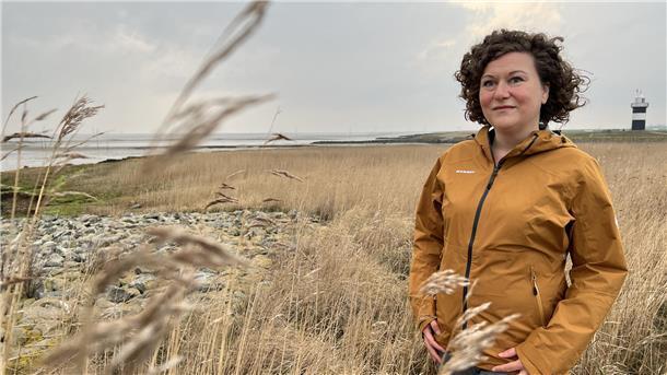 Frau mit braunen Locken steht an der Küste, im Hintergrund steht ein kleiner schwarz-weiß gestreifter Leuchtturm.
