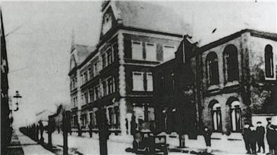 Ein altes Schwarz-Weiß-Foto zeigt ein historisches Gebäude mit mehreren Stockwerken und großen Fenstern. Im Vordergrund sind unscharfe Personen und ein Straßenlaternenpfahl erkennbar. Daneben steht ein weiteres, kleineres Gebäude.