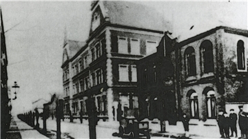 Ein altes Schwarz-Weiß-Foto zeigt ein historisches Gebäude mit mehreren Stockwerken und großen Fenstern. Im Vordergrund sind unscharfe Personen und ein Straßenlaternenpfahl erkennbar. Daneben steht ein weiteres, kleineres Gebäude.