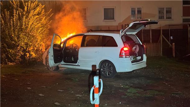 In einem Hof steht ein weißes Auto. Aus dem Motorraum schlagen Flammen.