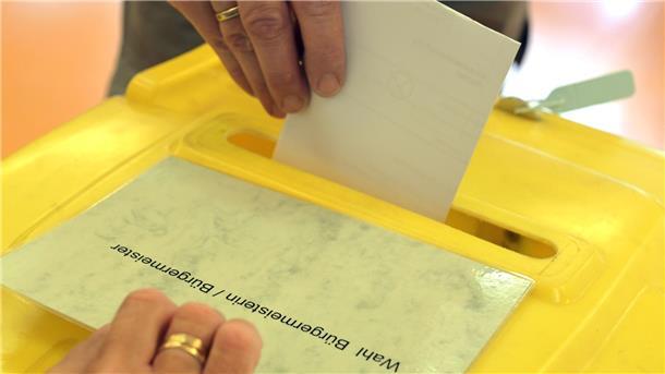 Eine Person wirft ihren Stimmzettel in eine gelbe Wahlurne. Auf dem Deckel steht „Bürgermeisterin / Bürgermeister“. Die Szene zeigt den Moment der Stimmabgabe bei einer Wahl.