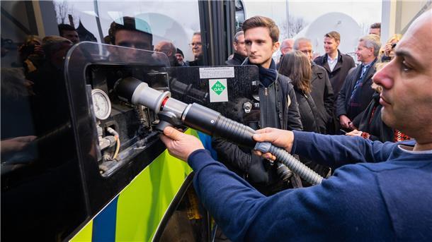Ein Mann betankt, umringt von Menschen, betankt einen Wasserstoffbus.