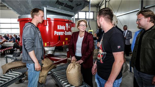 Niedersachsens Innenministerin Daniela Behrens steht mit mehreren Personen vor einer Sandsackfüllmaschine in einer Halle und hält einen gefüllten Sandsack.