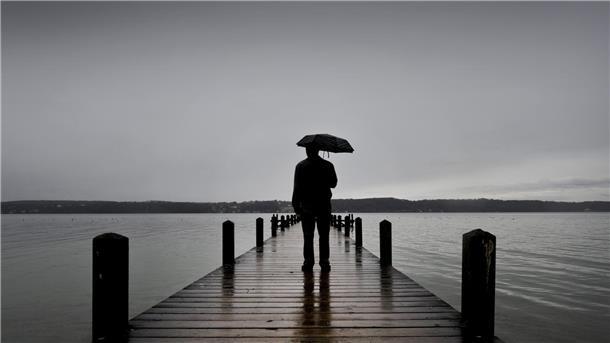 Ein Mann steht mit Regenschirm auf einem Steg. Man sieht nur seine Silhouette. Ringsherum ist Wasser. Der Himmel ist grau und wolkenverhangen.