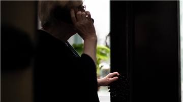 Eine Seniorin hält ein Telefon am Ohr und öffnet ihre Haustür einen Spalt weit.