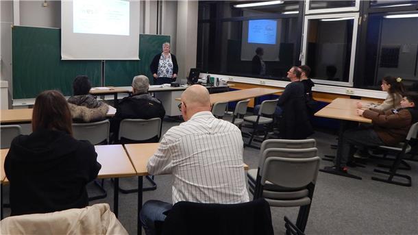 Mehrere Personen sitzen in einem Klassenzimmer und hören einer Frau zu, die vorne vor einer Tafel steht und eine Präsentation hält.