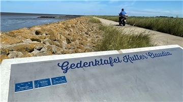 Die "Gedenktafel Kutter Claudia" an einer felsigen Küste. Im Hintergrund verläuft ein Strandweg, auf dem ein Mopdefahrer fährt.