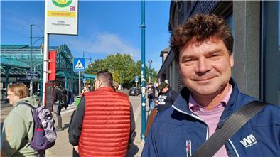 Ein Mann an einer Haltestelle blickt in die Kamera und lächelt.  