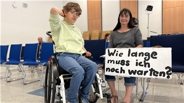 Junge im Rollstuhl, daneben seien Mutter. Sie hält ein Schild in der Hand. Darauf steht: "Wie lange muss ich noch warten?"