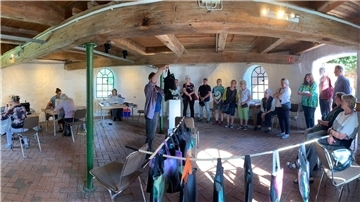 Kunstauktion in der Seefelder Mühle: Ein Mann hält eine Stofftasche zur Versteigerung hoch, im Hintergrund sitzen mehrere Frauen an Nähmaschinen, einige Besucherinnen und Besucher stehen ringsum.