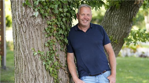 Ein Mann lehnt an einem Baum und schaut in die Kamera.